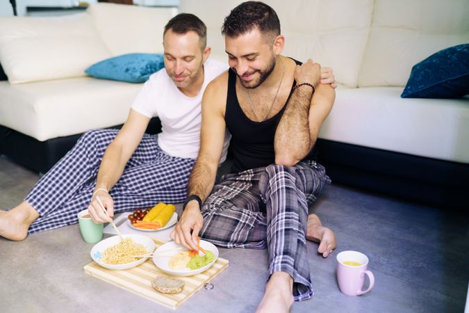 Happy male couple enjoying relaxing dinner at home sitting on floor