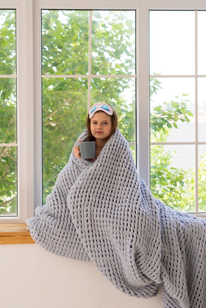 Girl wrapped in blanket by window with cup.