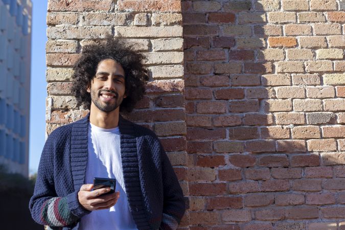 Happy man holding his smartphone while leaning on a brick wall outdoors on sunny day