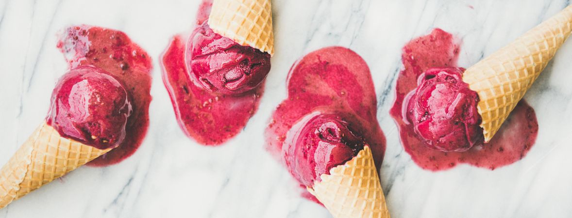 Four cones of dark berry ice cream melting on marble slab, horizontal composition