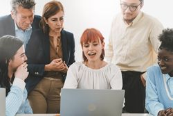 In a modern office setting, a redhead woman, sitting and explaining a presentation on her laptop to diverse colleagues