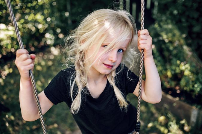Shy blonde girl with braided hair playing on outdoor swing in backyard