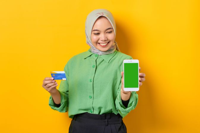 Happy Muslim woman in headscarf and green blouse holding credit card and smart phone screen mock up
