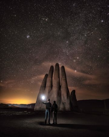 Silhouette of two people holding a torch standing beside hand shaped rock formation under starry night
