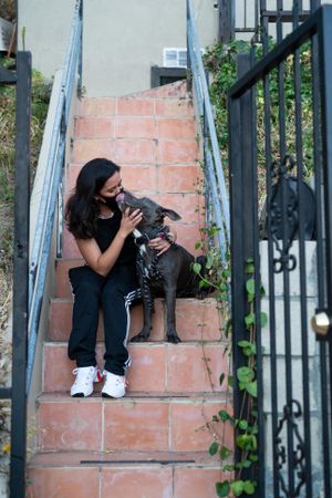 Woman receiving kiss from her dog while sitting on stairs in front of her house
