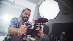 Young Male Videographer With Beard, Wearing Colorful Shirt And Cap, Enthusiastically Operates Camera In Studio Setting, Capturing Content For Advertising Or Movie Production.
