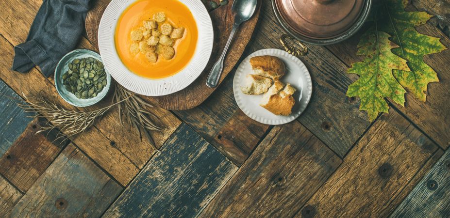 Pumpkin soup with croutons, bread, seeds, pot, spoon on wooden table, wide composition, copy space
