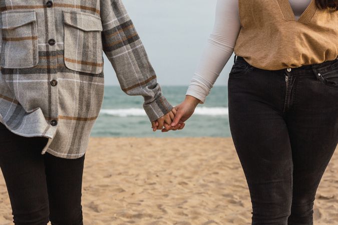 Crop of women holding hands while walking on sandy seacoast