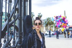Front view of a young laughing hipster woman standing and leaning on a fence at park in sunny day while looking away