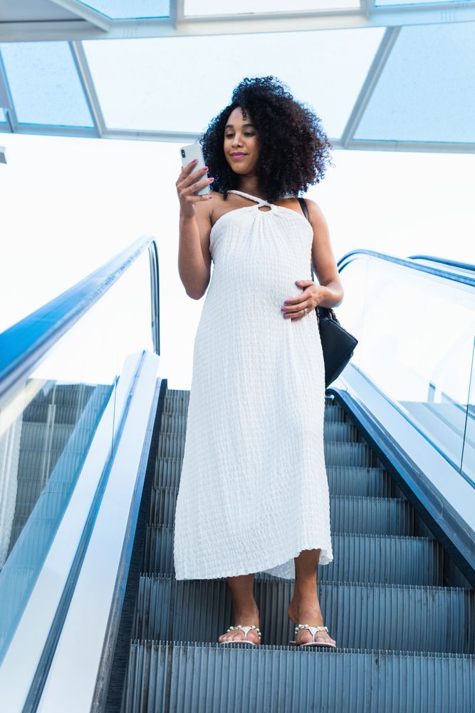 Pregnant Black woman browsing smartphone on escalator