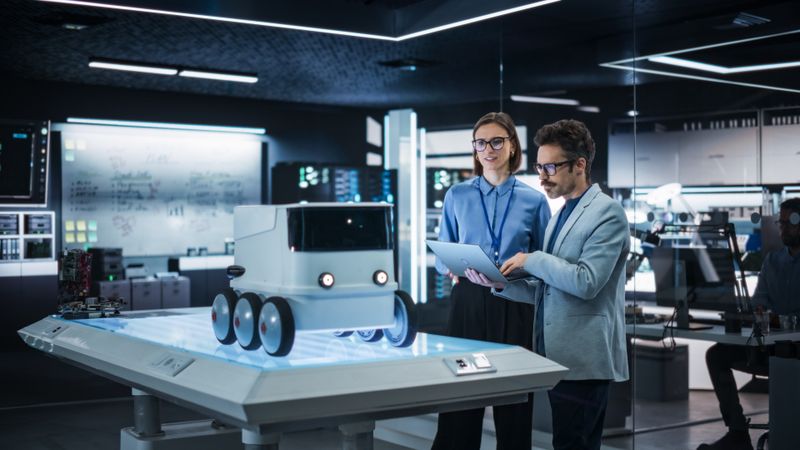Portrait of Two Colleagues Working in a Research Facility, Using Laptop Computer to Develop a Futuristic Robot for Autonomous Deliveries. Manufacturing Specialist Doing a Demonstration to Team Manager