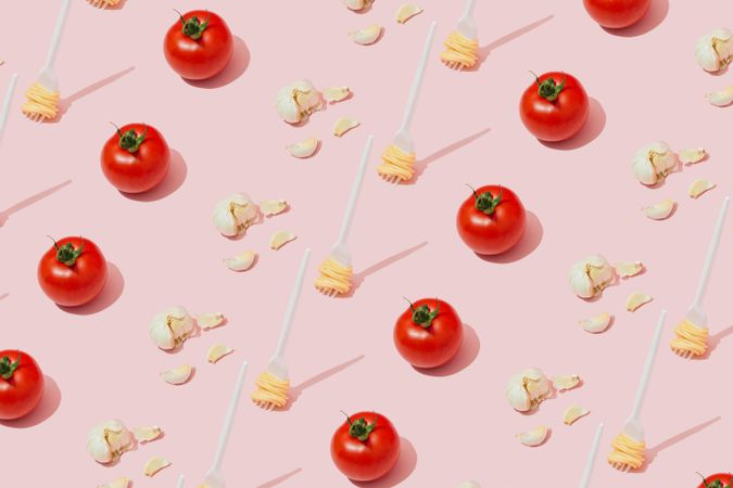 Rows of garlic bulbs, tomatoes, and plastic fork with pasta pattern against pastel pink background