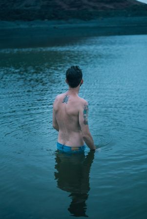 Back of tattooed male standing in water with jeans
