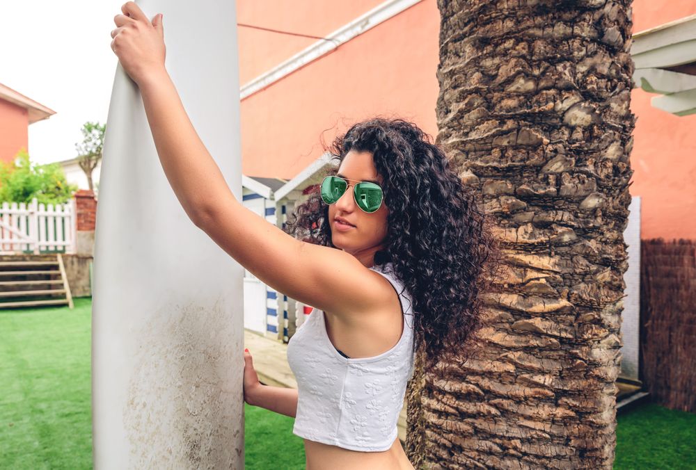 Brunette surfer woman in sunglasses and bikini holding surfboard next to palm tree
