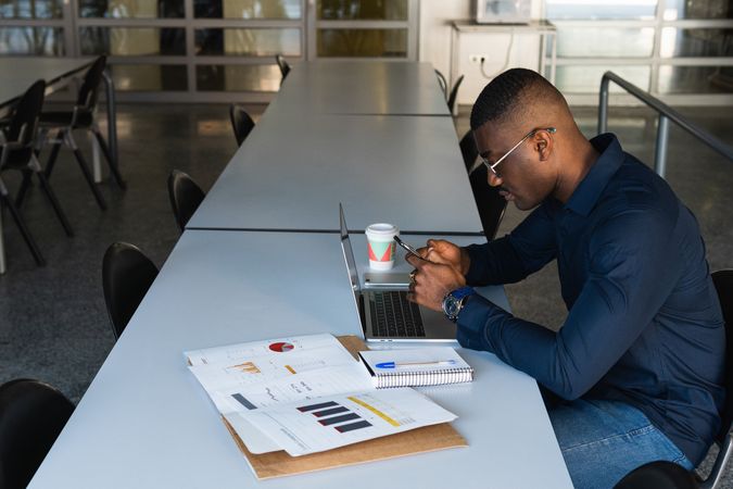 Black man working on laptop with smartphone in office