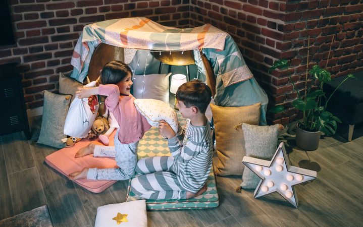 Excited boy and girl laughing and playing in cushion battle on teepee