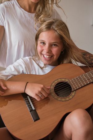 Portrait of a blonde girl playing guitar while smiling on camera with a woman sitting with he