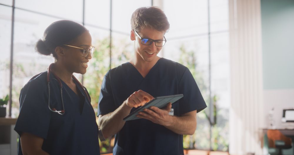 Two Young Doctors Standing in a Medical Facility in the Hospital, Talking and Sharing Information on Tablet Computer.