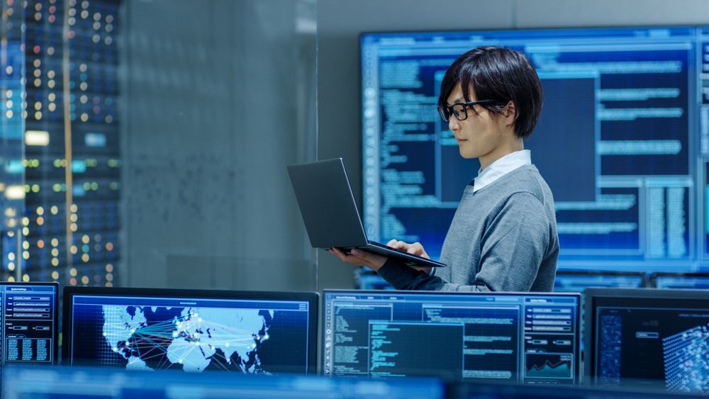 In the System Control Room IT Technician Works on a Laptop, in Background Multiple Displays with Graphics.