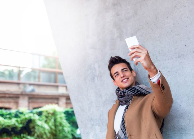 Smiling male in camel coat and scarf standing outside taking selfie