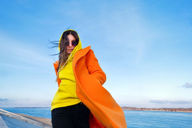Woman in sunglasses, orange coat, yellow hoodie and jeans against the bay background