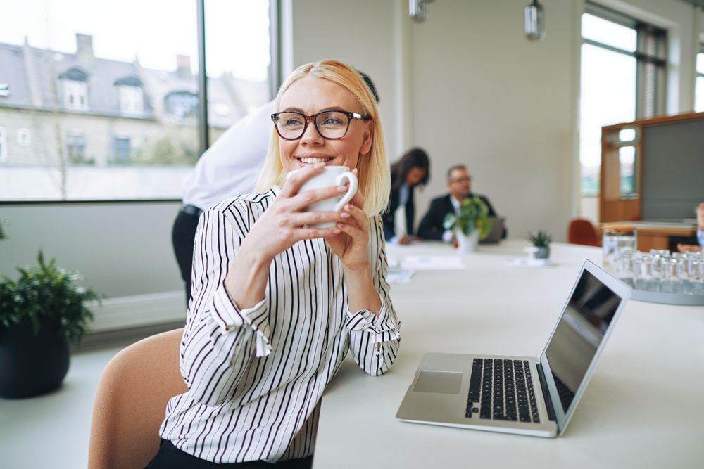 Woman looking up from her laptop and smiling in a bright office drinking coffee