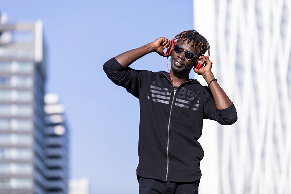 Happy Black man wearing headphones & sunglasses standing on the street listening to music