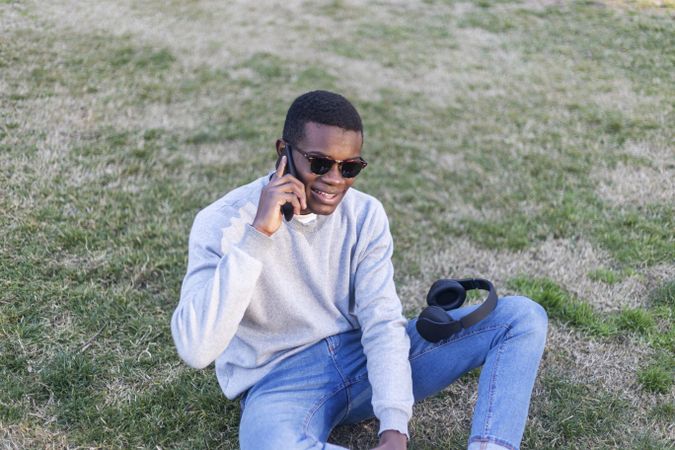Happy Black male with sunglasses sitting in the grass talking on his phone