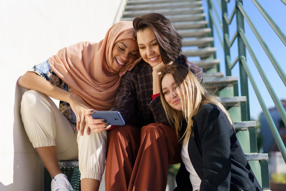 Multi-ethnic women friends in stylish outfits looking at phone on metal stairs outside
