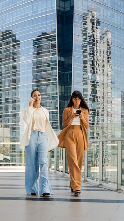 Two women are walking on a sidewalk, one of them talking on a cell phone and the other woman is writing a message. The scene is set in a city with a tall building in the background.