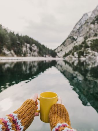 Person with cup near wonderful lake between hills in snow and cloudy sky