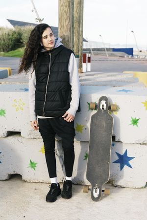 Teenage girl leaning against a wall next to her longboard while looking away from camera