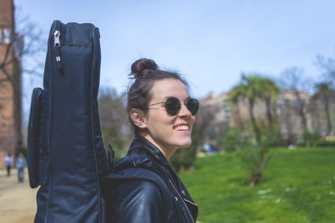 Side view of a young smiling hipster woman walking in a park in sunny day while carrying a guitar on back