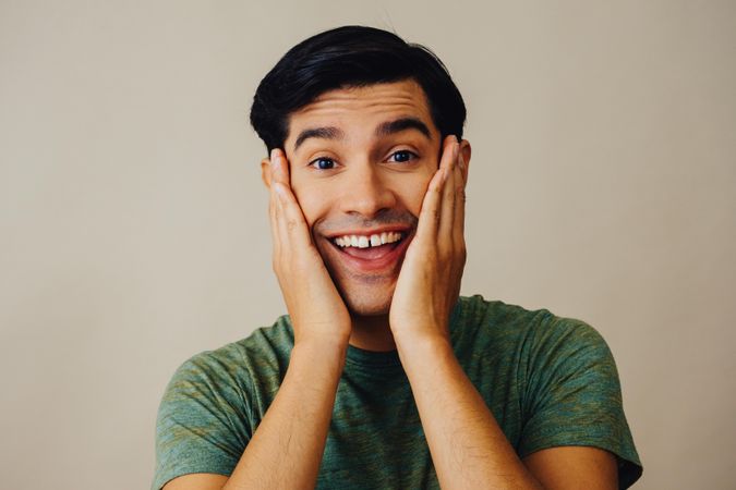 Portrait of Hispanic male in neutral room with hands on both cheeks