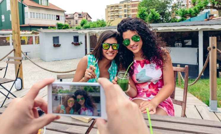 Two women sipping cocktails outside as friend takes photo