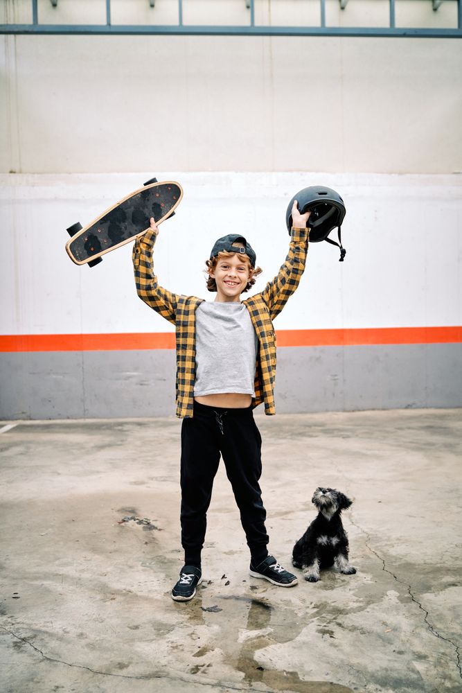 Cheerful child raising skateboard with helmet near dog on street