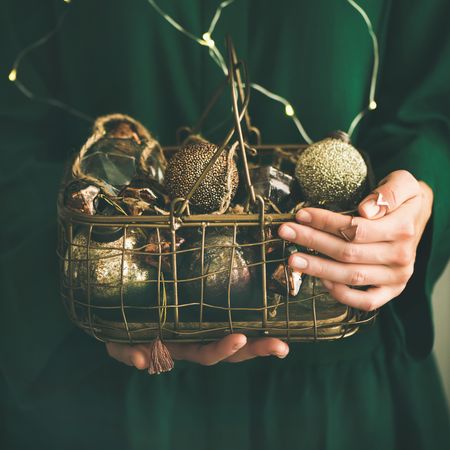 Woman in green dress holding wire basket of gold holiday decorations, square crop