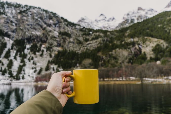 Person with cup near wonderful lake between hills in snow and cloudy sky