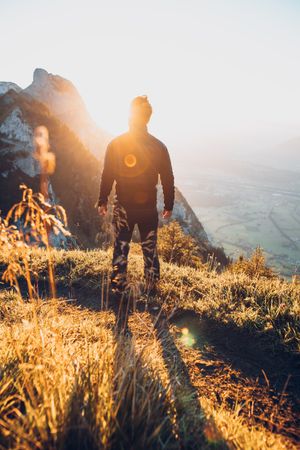 Silhouette of a man standing on brown grass facing the sun and mountains