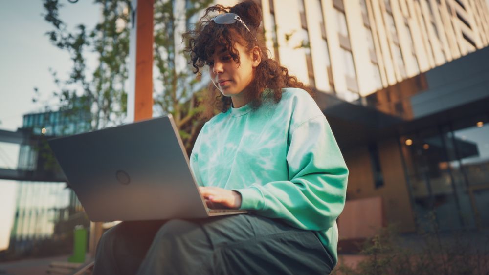 Young Woman With Curly Hair, Wearing A Tie-dye Sweatshirt, Works Intently On A Laptop Outdoors