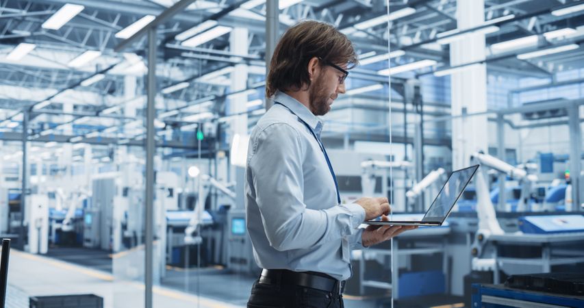 Experienced Male Engineer Standing in Office, Using Laptop Computer and Overlooking Production at Modern Automated Electronics Assembly Line with AI Support. Factory Executive at Work