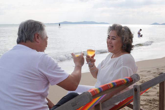 Happy mature Asian couple relaxing on the beach and toasting with wine