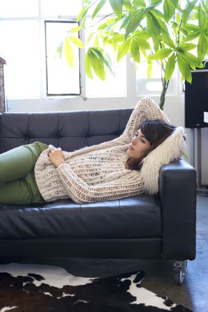 Female in wooly sweater laying on her side trying to nap on leather couch in living room, vertical
