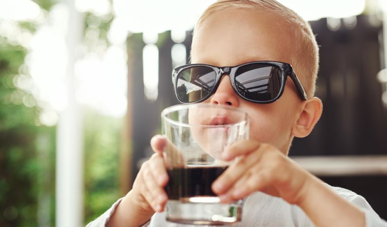 Blond boy in sunglasses sipping dark drink from glass outside