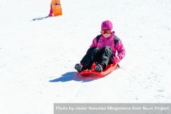 Child in pink snow suit going down a snowy hill in a plastic sled 0J1kdb