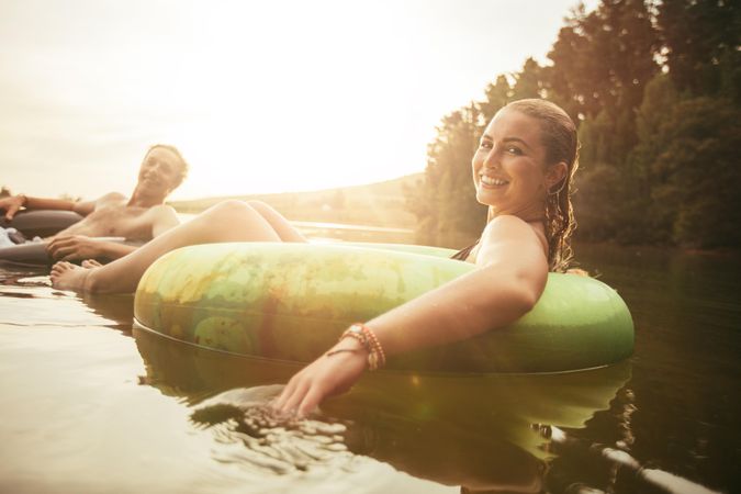 Portrait of happy young woman in lake on inflatable ring with her boyfriend in the background