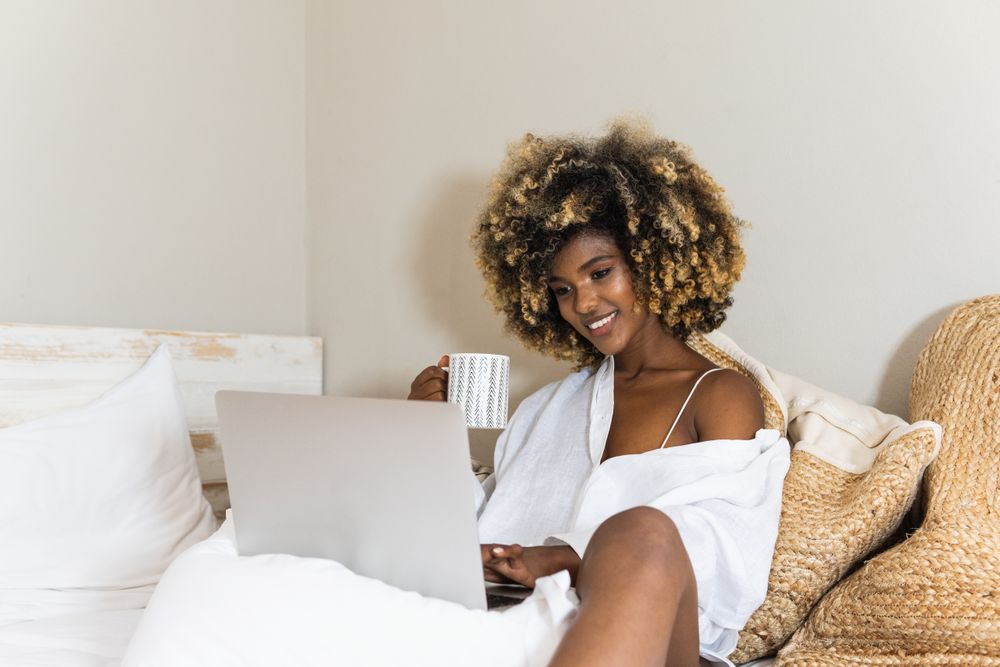 Black woman drinking coffee while sitting on bed with laptop