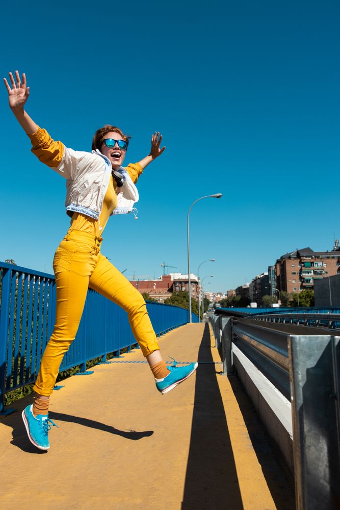 Stylish young carefree woman in vibrant clothes having fun on street bridge against blue sky