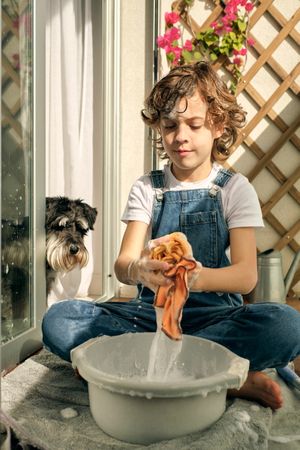 Child wringing out rag near dog while washing balcony