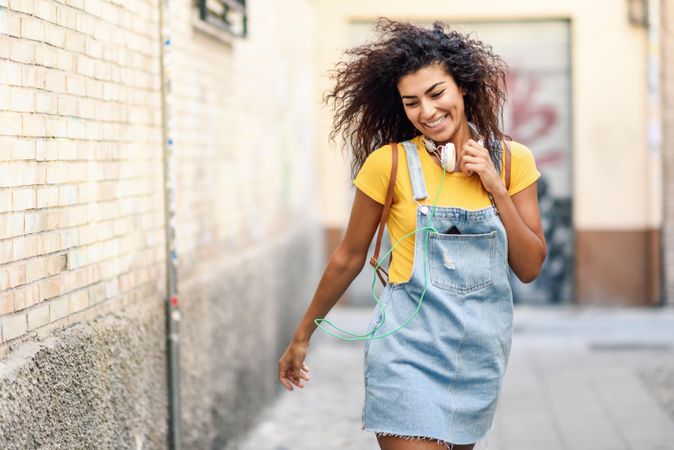 Dancing Arab woman with headphones and curly hairstyle walking outdoors in lane with graffiti